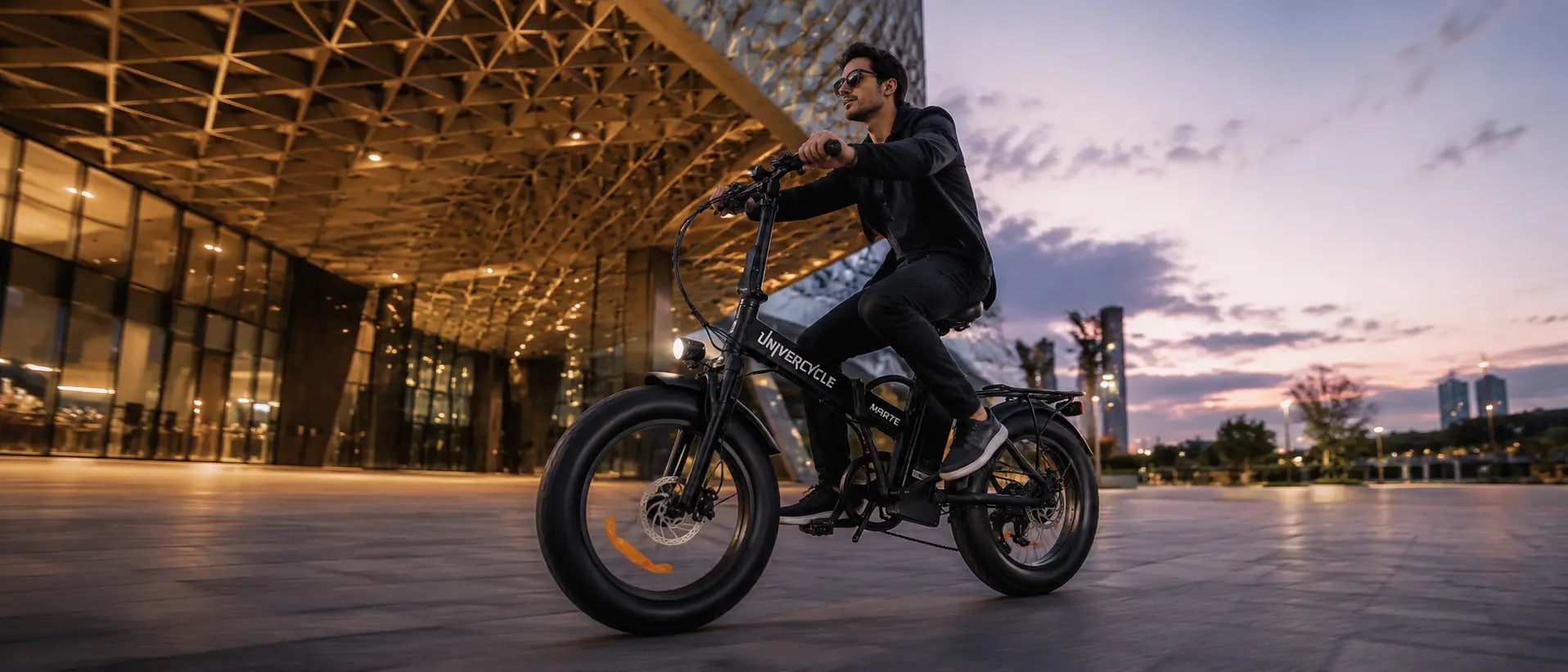 Desktop horizontal banner showing a man riding the black UNIVERCYCLE Marte folding electric fat bike at twilight, with an illuminated modern geometric building and sunset sky in the background.
