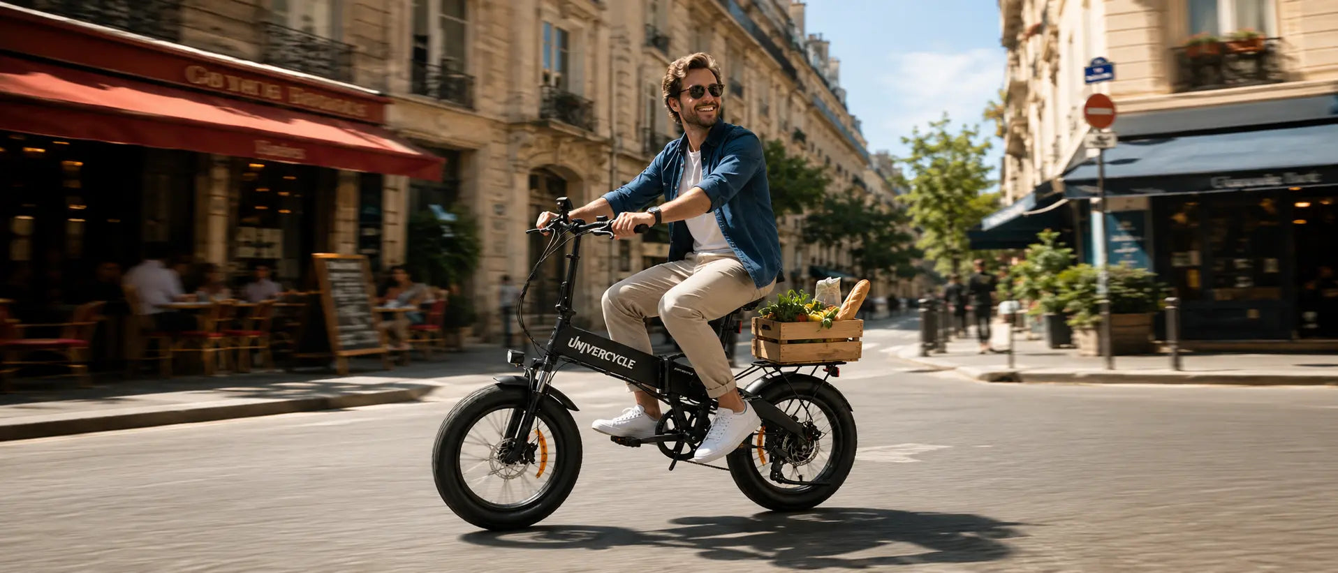 A man wearing sunglasses riding the UNIVERCYCLE Saturno folding electric bike on a European city street, with a wooden crate filled with groceries on the rear rack.